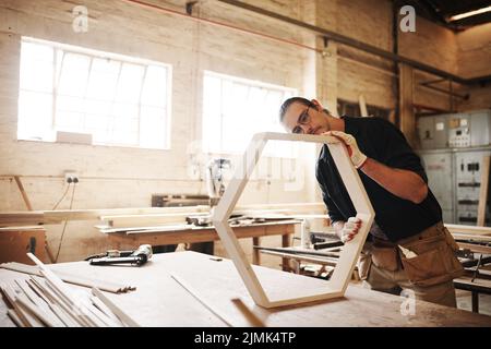 Die Liebe zum Detail ist in dieser Branche von entscheidender Bedeutung. Ein hübscher junger Zimmermann, der in einer Werkstatt an einem Holzrahmen arbeitet. Stockfoto