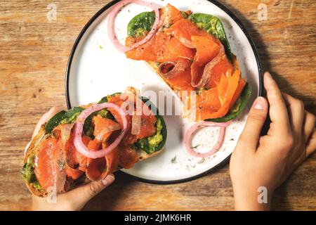 Weibliche Hände und leckere Toasts mit geräuchertem Lachs, Avocado und Basilikumblättern Stockfoto