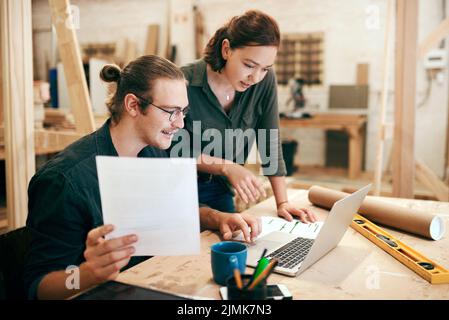 Zwei junge Zimmerleute, die in ihrer Werkstatt zusammen an einem Laptop arbeiten und planen, arbeiteten an etwas bedeutendem für unser Geschäft. Stockfoto