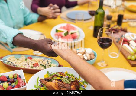 Mehrrassische ältere männliche und weibliche Freunde halten die Hände und beten am Tisch Stockfoto