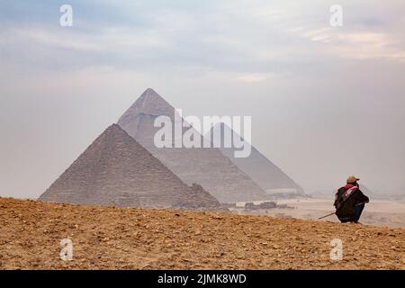 Ägyptischer Mann, der auf einem Hügel mit den verschwommenen Pyramiden im Hintergrund sitzt, Kairo, Ägypten. Stockfoto
