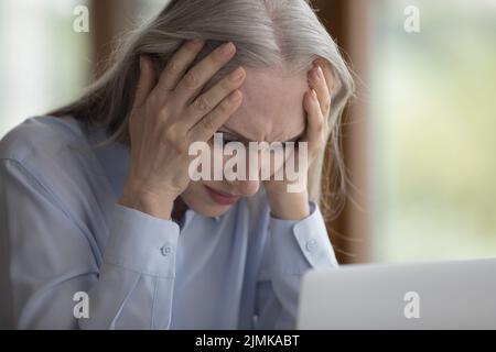 Schockiert frustrierte ältere Frau, die den Kopf auf den Laptop berührte Stockfoto