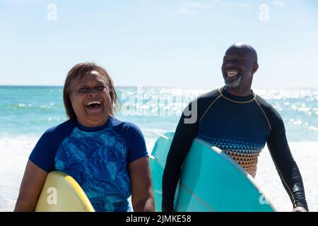 Fröhliches afroamerikanisches Senioren-Rentnerpaar mit Surfbrettern, das einen sonnigen Tag am Strand genießt Stockfoto