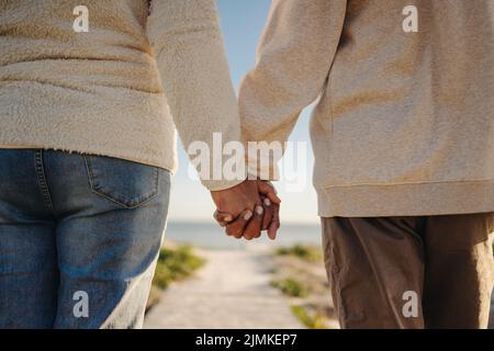 Romantisches Seniorenpaar, das sich die Hände hält, während es am Strand eine Strandpromenade entlang geht. Nicht erkennbares älteres Paar, das einen erfrischenden Badeurlaub af Stockfoto