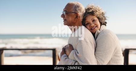 Romantische ältere Frau lächelt der Kamera zu, während sie ihren Mann am Meer umarmt. Liebevolles, älteres Paar genießt es, etwas Zeit zu verbringen Stockfoto