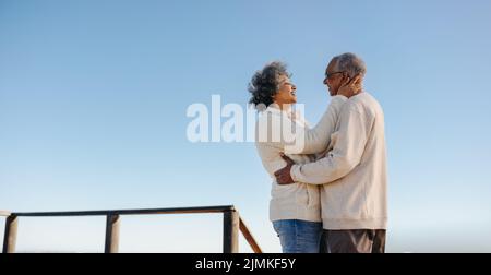 Ein glückliches Seniorenpaar lächelt sich gegenseitig an, während es auf einer Promenade am Strand steht. Ein romantisches, älteres Paar genießt es, eine gute Zeit zusammen zu verbringen Stockfoto