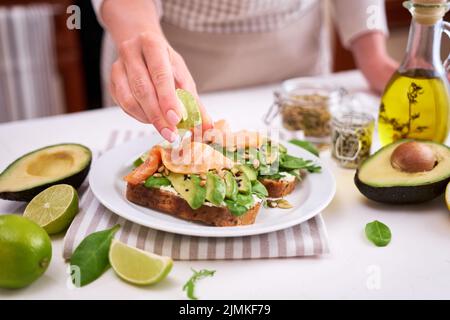 Frau quetscht Limettensaft auf frisch zubereiteten Avocado-, Lachs- und Frischkäse-Toasts Stockfoto