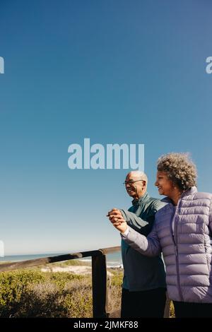 Romantischer Strandspaziergang. Rentnerehepaar lächelt und hält sich die Hände, während sie zusammen auf einer Promenade spazieren geht. Glückliches älteres Paar enjoyi Stockfoto