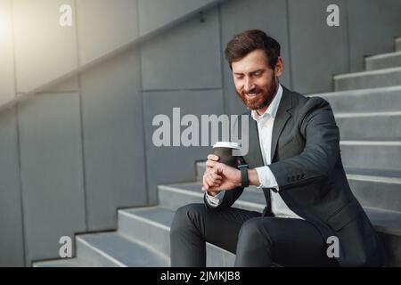 Attraktiver Büroangestellter im Anzug, der während der Pause auf der Treppe sitzt und Kaffee trinken und auf seine Uhr schaut Stockfoto