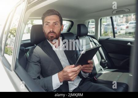 Ein gutaussehender Geschäftsmann, der in einem Luxusauto auf einem digitalen Tablet sitzt Stockfoto