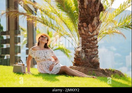 Ein helles schönes Mädchen in einem hellen Kleid und Hut sitzt auf dem Gras unter einer Palme in Monaco bei sonnigem Wetter im Sommer, stroh Stockfoto