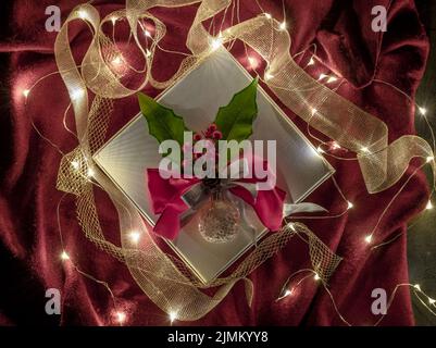 Ein Weihnachtsgeschenk ist mit goldenen Bändern, grünem und rotem Stechpalme und glühenden Lichterketten auf rotem Hintergrund dekoriert Stockfoto