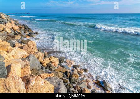 Wellen schlagen auf der Ufermauer mit großen Felsen in San Clemente, Kalifornien. Meer mit Blick auf den Pier auf der Rückseite gegen den klaren blauen Himmel. Stockfoto