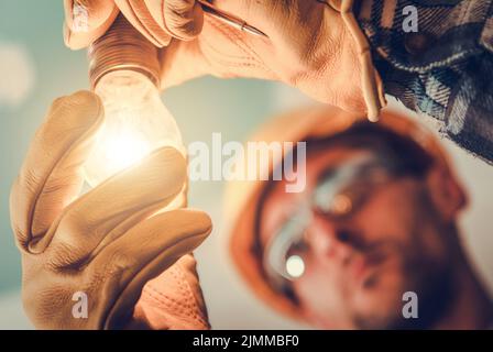 Nahaufnahme eines kaukasischen Elektrikers, der einen Harthut und Schutzhandschuhe trägt und einen leuchtenden Lichtpunkt in seinen Händen hält. Installation Der Beleuchtung Erfolgreich Stockfoto