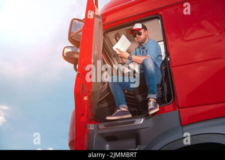 Kaukasischer Lastwagenfahrer, der in der Kabine seines roten Sattelwagens sitzt, überprüft sorgfältig die Transitpapiere, bevor er eine Route mit einem großen Frachtgut anlegt Stockfoto