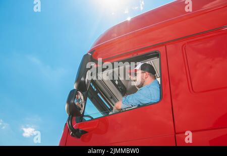 LKW-Fahrer in einem blauen Hemd und Baseballmütze, der in seinem riesigen Reg-Lastwagen sitzt und die Straße vor sich sieht. Blauer sonniger Himmel im Hintergrund. Transportation Stockfoto