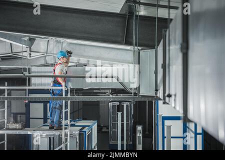 Kaukasischer Arbeiter trägt Sicherheitsuniform und blaue Hartmütze Überprüfung mit einer Fackel das Belüftungssystem in riesigen kommerziellen Halle. Stockfoto