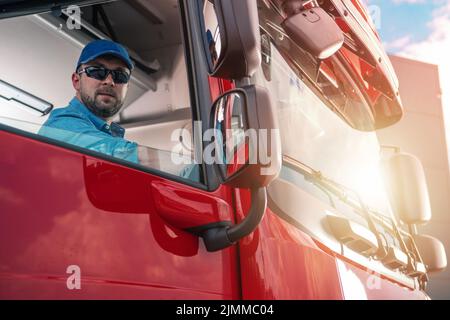 Kaukasischer Trucker in seinem 30s sitzenden Passagier in der Sear eines roten Semi-Trucks, der aus einem offenen Fenster blickt. Ruhe während einer langen Fahrt. Transportmittel Stockfoto