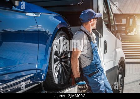 Kaukasischer Fahrer mittleren Alters, der auf der Seite seines Abschleppwagens sitzt, beladen mit dem Auto, das in Geduld auf die Freigabe wartet, um den neuen zu liefern Stockfoto