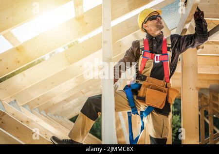 Kaukasischer Auftragnehmer, der Sicherheitskabelbaum und Schutzhelm trägt, überprüft die Festigkeit der Installation von Holzbohlen im Dachabschnitt eines Holzresims Stockfoto