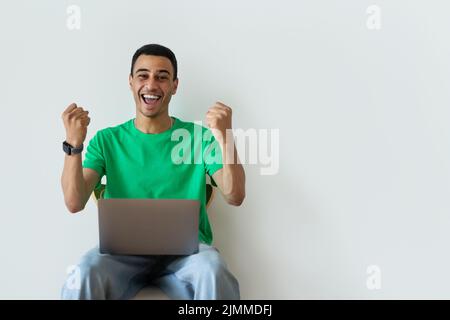 Überfröhlicher araber, der mit JA-Geste auf einem Stuhl mit Laptop sitzt und über einer weißen Wand Erfolg oder Erfolg feiert Stockfoto