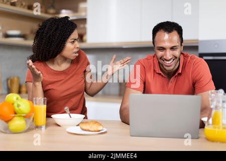 Enttäuscht wütend junge afroamerikanische Frau schreit auf lächelnden Mann, Kerl spielen auf Laptop Stockfoto