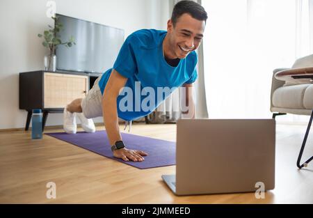 Glücklicher arabischer Mann in Sportswear, der Liegestütze macht, Laptop benutzt, von zu Hause aus einen Online-Fitnesskurs macht, Wohnzimmerinnenraum nutzt Stockfoto
