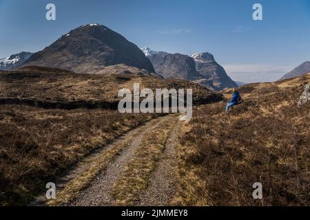 Touristen, die neben der alten Militärstraße saßen und auf den Schnee schauten, überstiegen Stob Coire nan Lochan und die drei Schwestern unter einem klaren blauen Himmel. Stockfoto