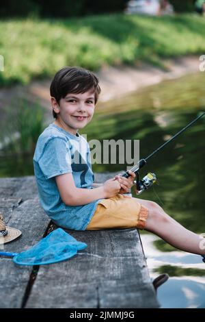 Glücklicher Junge sitzt hölzernen Pier See hält Angelrute Stockfoto