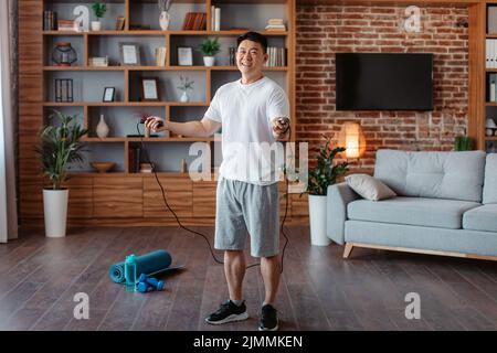 Sportlicher Mann mittleren Alters mit Springseil, der zu Hause trainiert, häusliches Training im Wohnzimmer, in voller Länge Stockfoto