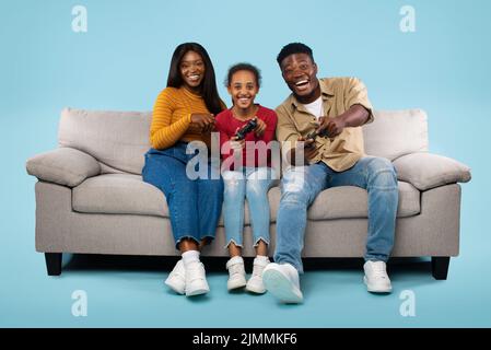 Überglücklich afroamerikanische Eltern und ihre Tochter spielen Videospiele und sitzen auf dem Sofa auf blauem Hintergrund Stockfoto