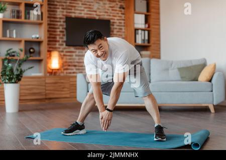 Aktiver asiatischer reifer Mann, der Übungen macht, die Beinmuskeln zu Hause auf der Fitnessmatte dehnt, in voller Länge, Platz kopieren Stockfoto