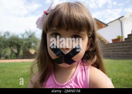 Bild eines entzückenden kleinen Mädchens mit einem traurigen Ausdruck, das ihren Mund zugeklebt hat. Stockfoto