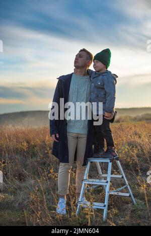 Kaukasischer Vater steht mit seinem Sohn auf dem Hügel und schaut weit weg Sonnenuntergang Herbst Zeit Stockfoto