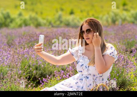 Hübsche junge Frau, die ein Selfie in einem Lavendelfeld macht, während sie im Sommer entspannt zwischen den Blumen sitzt Stockfoto