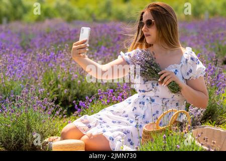 Junge Frau, die ein Selfie macht, während sie sich auf einem Bauernhof zwischen Lavendelfeldern entspannt und einen Strauß frischer Blumen an ihre Brust hält Stockfoto