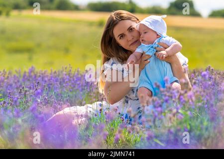 Entzückender kleiner Junge in einem blauen Jumpsuit, der von seiner liebevollen Mutter gehalten wird, die sich zwischen violetten Lavendelblüten entspannt Stockfoto