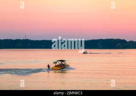 Ein Boot zieht eine Person auf einem Wakeboard über die Wellen. Wakeboarding bei Sonnenuntergang. Stockfoto