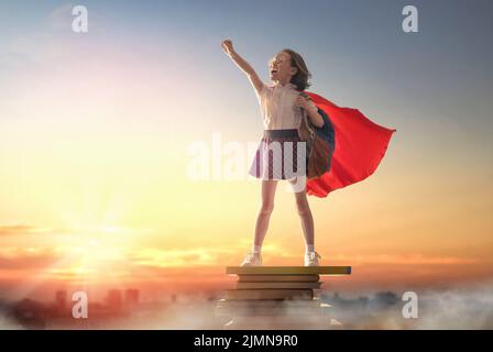 Zurück zur Schule! Happy cute fleißig Kind auf dem Turm von Büchern auf dem Hintergrund des Sonnenuntergangs Himmel. Mädchen trägt Superhelden Umhang. Konzept von su Stockfoto