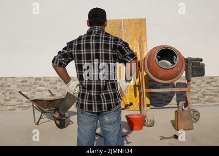 Bauarbeiter mit Hammer und Kelle auf einer Baustelle. Hintergrund eine Schubkarre und ein Betonmischer mit Beton. Stockfoto