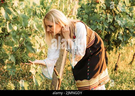 Junge blonde Frau in serbischer traditioneller Kleidung mit Blick auf grüne Trauben Stockfoto