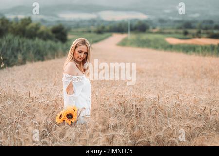 Junge, blonde Frau, die auf dem Weizenfeld mit Sonnenblumen in der Hand steht Stockfoto