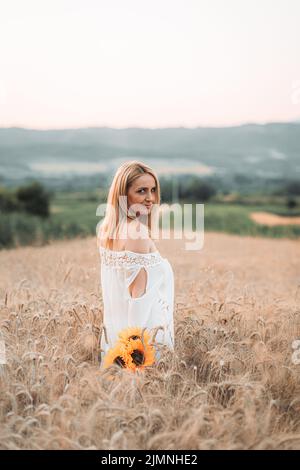 Junge, blonde Frau, die auf dem Weizenfeld mit Sonnenblumen in der Hand steht Stockfoto