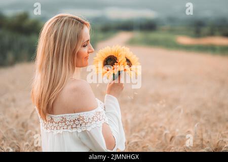 Junge, blonde Frau, die auf dem Weizenfeld mit Sonnenblumen in der Hand steht. Speicherplatz kopieren Stockfoto