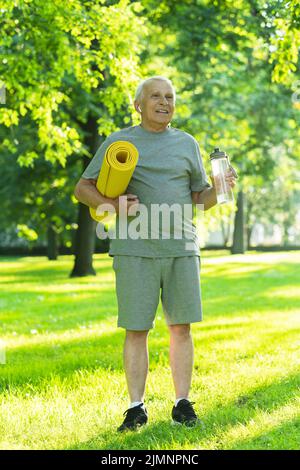 Aktiver älterer Mann mit einer Trainingsmatte und einer Flasche Wasser im grünen Stadtpark Stockfoto