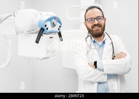 Lächelnder, selbstbewusster Radiologe und Stehen in der Nähe von Röntgengeräten. Stockfoto