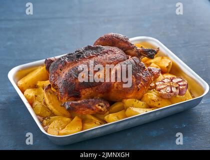 Gebackenes Hähnchen in blauer Auflaufform auf dunkelblauem Tisch, gebratenes Fleisch mit Kartoffeln. Seitenansicht Stockfoto