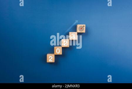 2023-Jahres-Zahlen auf Holzblöcken stapeln, Diagrammschritte mit leuchtendem Aufwärtspfeil zum Zielsymbol auf blauem Hintergrund, Geschäftsprozess bis Stockfoto