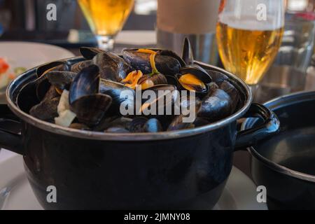 Gekochte Muscheln in einer schwarzen Schüssel und Bier in einem Restaurant. Meeresfrüchte. Klassische traditionelle französische Küche. Leckeres Abendessen. Stockfoto