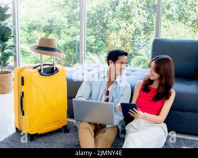 Glückliches asiatisches Paar, junger Mann und Frau, die sich gegenseitig suchen, während sie Laptop und Tablet zusammen für Flugbuchungen und Reiseinformationen in verwenden Stockfoto